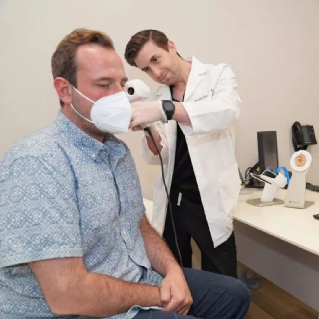 The image shows a person sitting in a dental chair receiving treatment from a dentist wearing a face mask and stethoscope, with a man in a lab coat adjusting the mask on the seated individual s face.