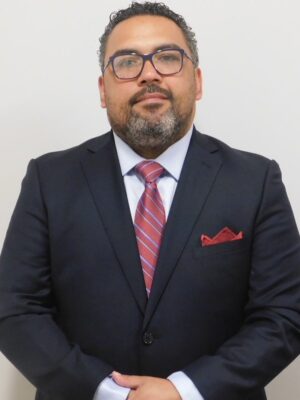 The image shows a man wearing a suit with a red tie, standing against a white background, with his arms crossed and hands in pockets. He has glasses and appears to be balding, with a beard.